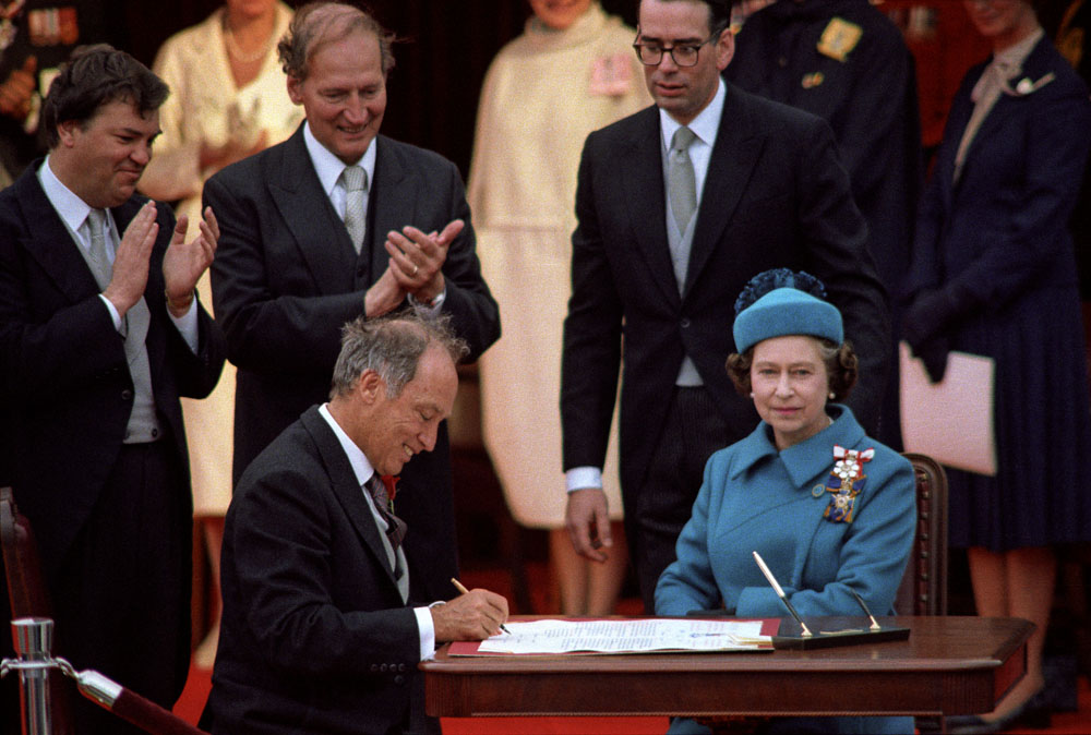 Prime Minister, Pierre Elliott Trudeau sitting at a table with Queen Elizabeth II as he signs the Proclamation of the Constitution Act, 1982.